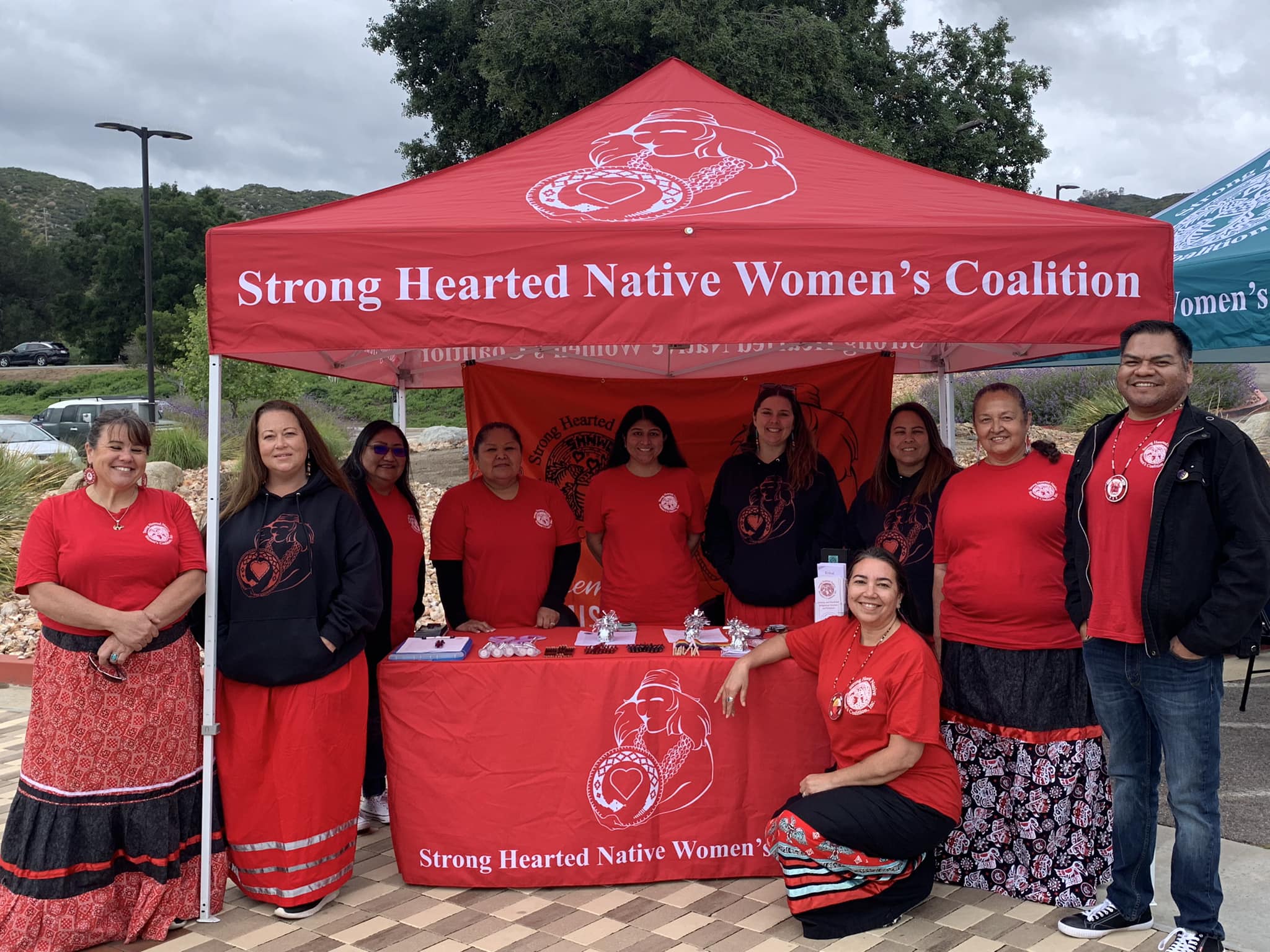 standing and sitting at the Strong Hearted Native Women’s Coalition booth under a red tent with a banner. They are wearing red shirts and traditional ribbon skirts standing and sitting at the Strong Hearted Native Women’s Coalition booth under a red tent with a banner. They are wearing red shirts and traditional ribbon skirts.