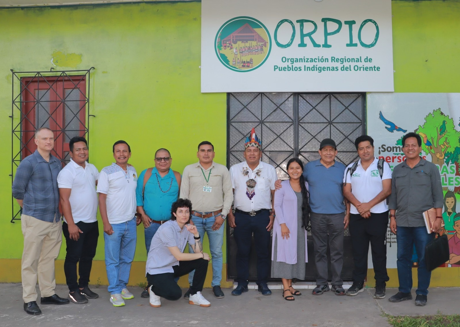 Group of ten people smiling and standing outside the office of ORPIO, Organización Regional de Pueblos Indígenas del Oriente, under its signboard.