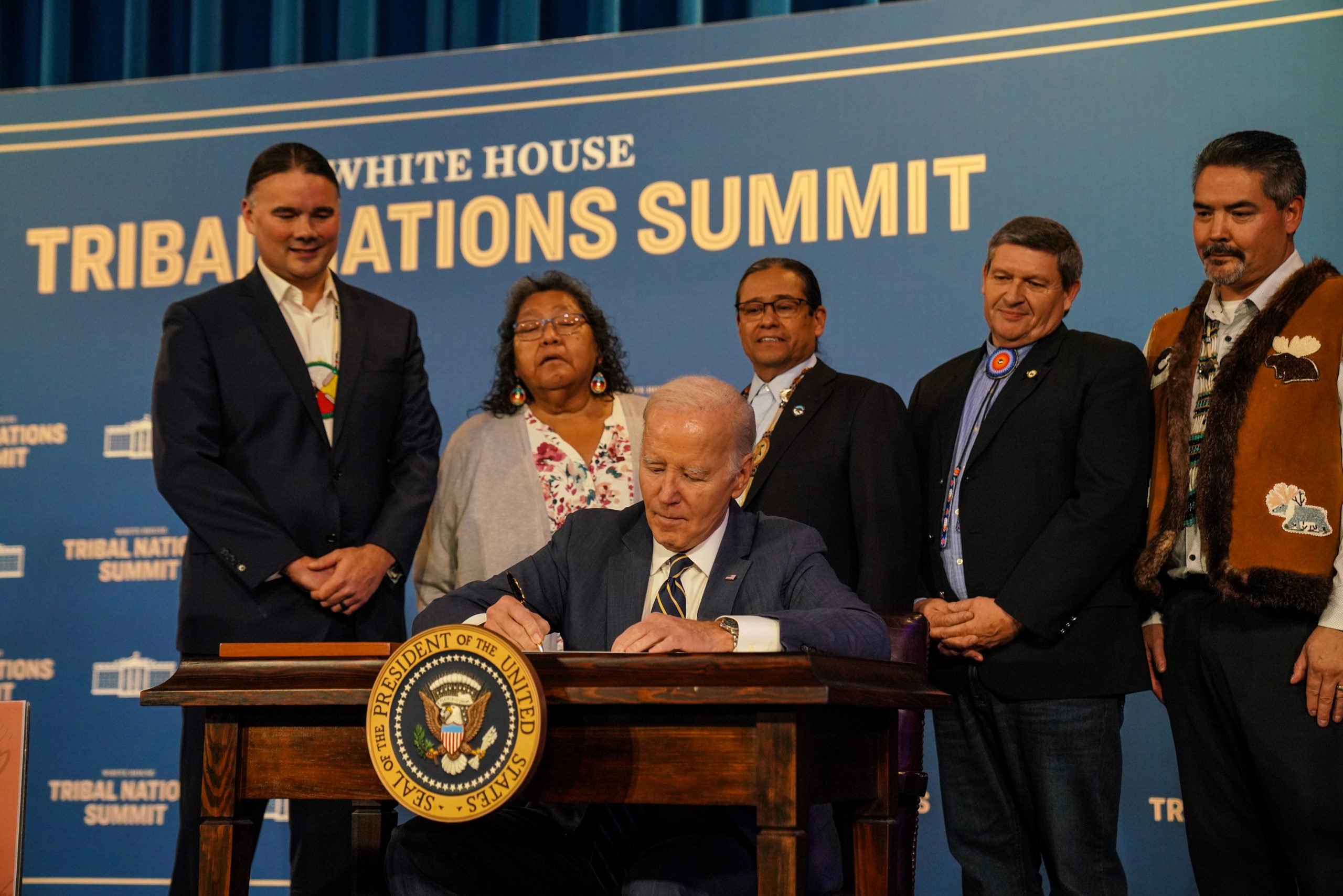 President Joe Biden signing a document at the Tribal Nations Summit, flanked by five attendees in formal attire.