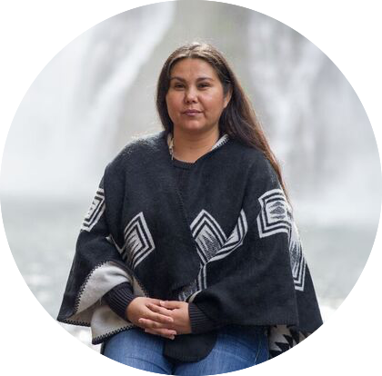 A woman with long dark hair sits outdoors in front of a waterfall, wearing a dark poncho with geometric patterns. She is looking calmly into the camera, hands resting in her lap, with a serene and natural background of flowing water and soft light.