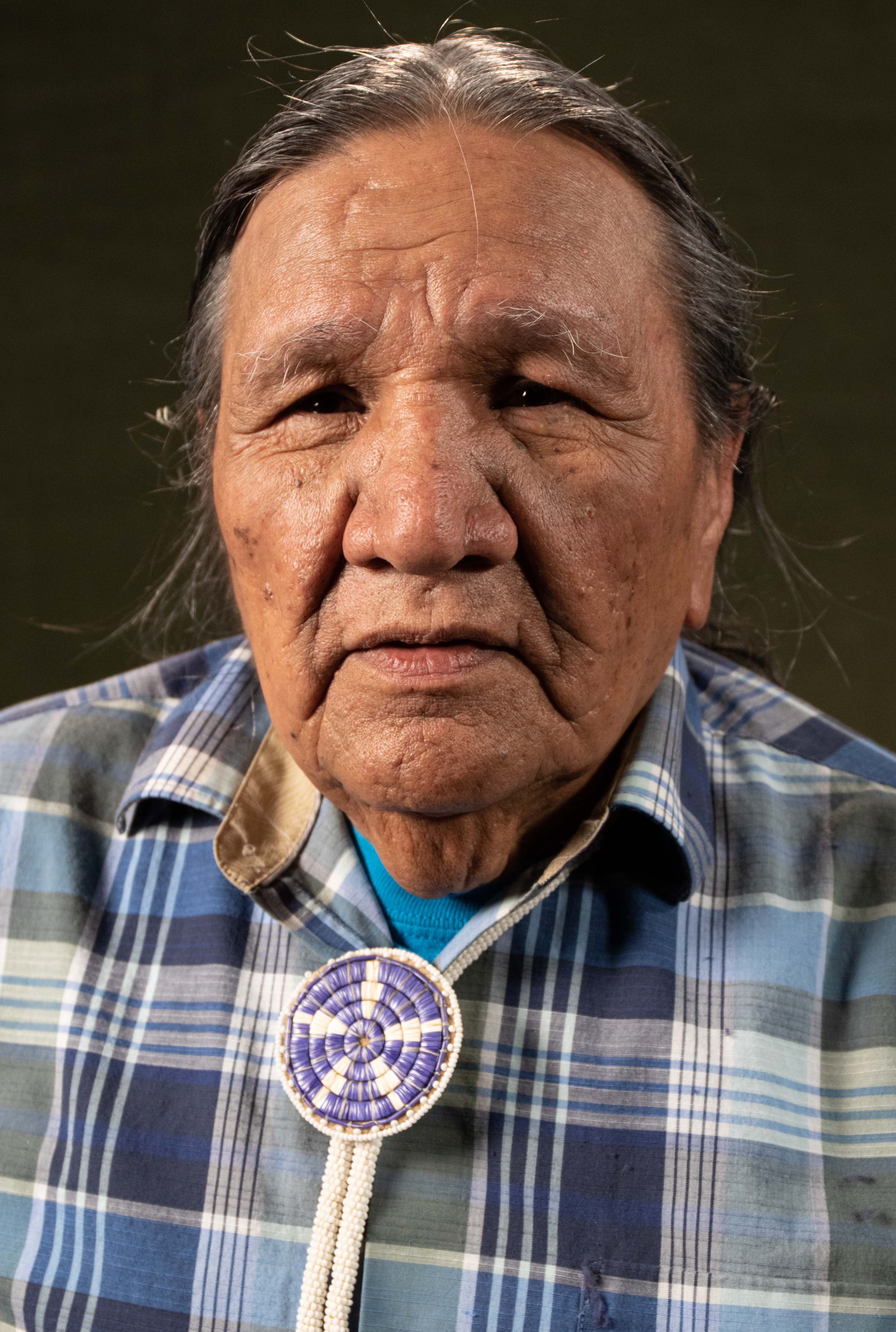 Portrait of an elder wearing a plaid shirt and a beaded medallion, with a thoughtful expression.