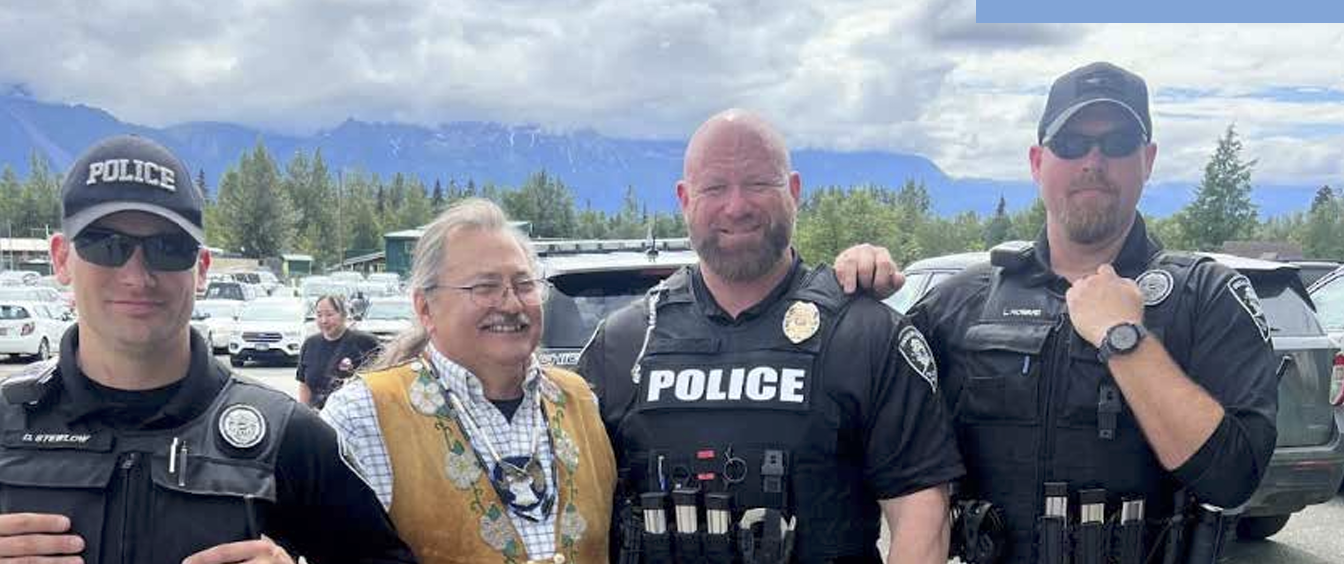 Four individuals, including one smiling in traditional indigenous attire, standing together outdoors with two police vehicles in the background, under a cloudy sky with mountains.