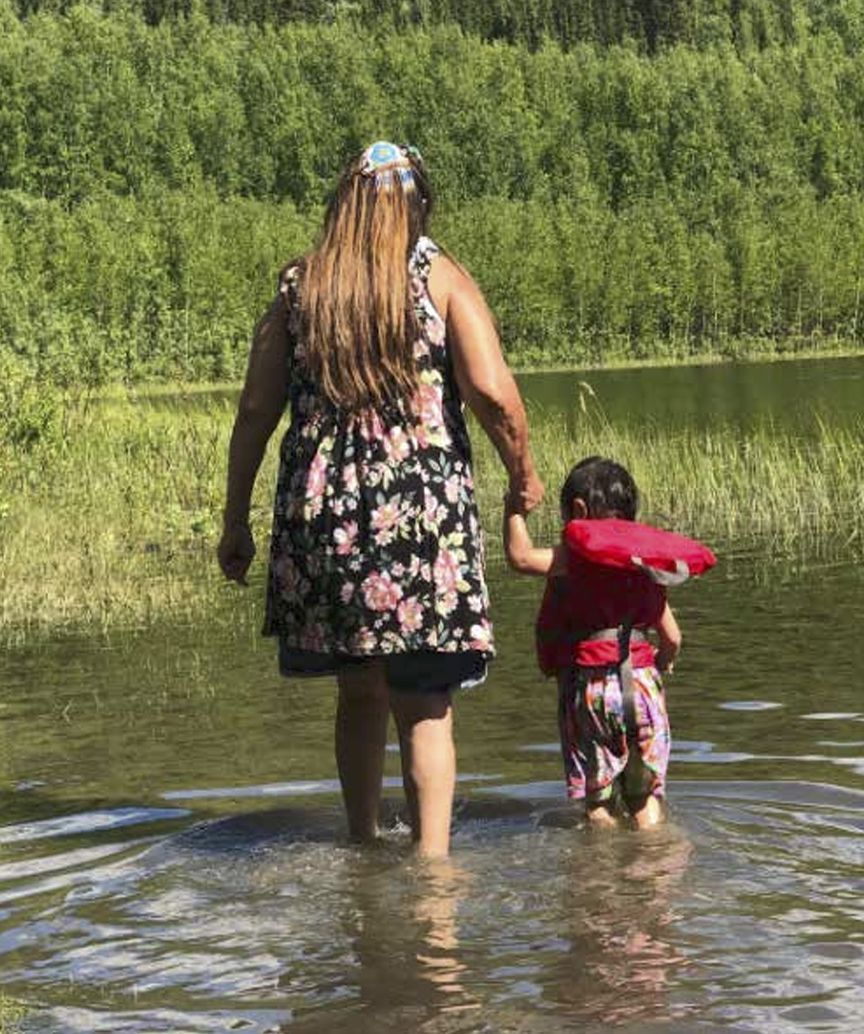 Two people, an adult and a child, wading into a lake surrounded by greenery on a sunny day. The adult is wearing a floral dress and the child is in colorful attire with a red life jacket.