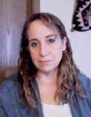 Rebekah Jones with medium-length curly hair wearing a denim jacket, sitting in front of a wooden background with a decorative textile hanging.