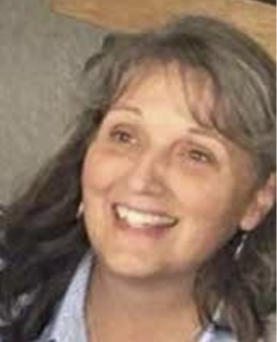 Portrait of Mary Mummaw with shoulder-length wavy hair, wearing a denim jacket. They are indoors with a wooden background.