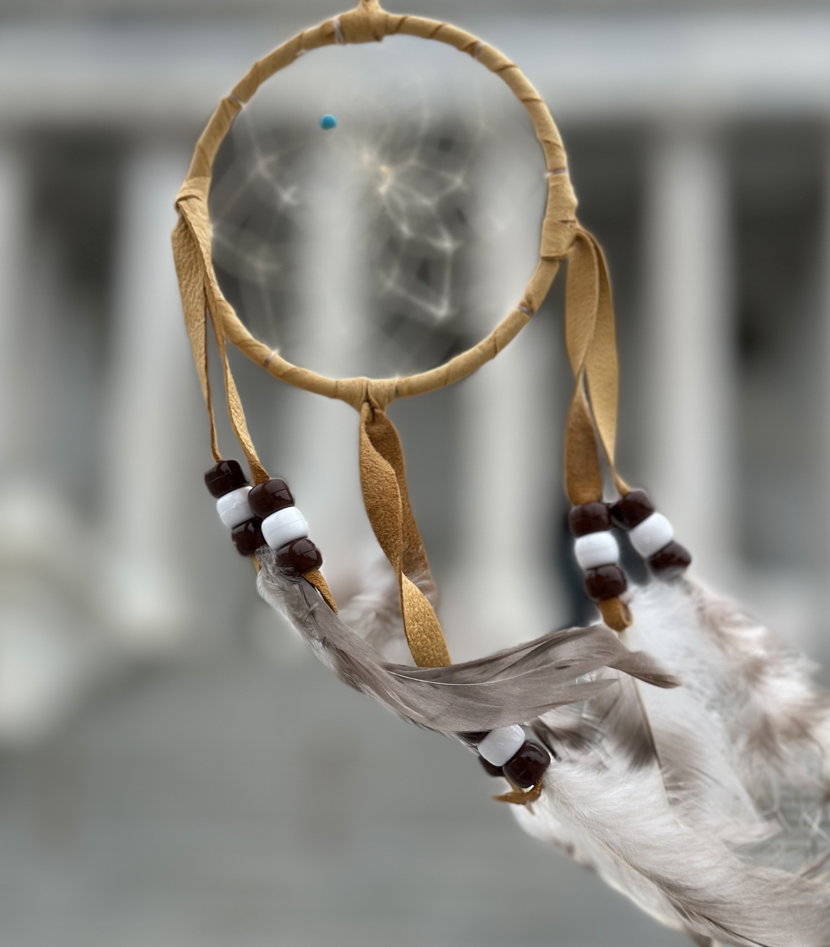 A close-up of a dreamcatcher with white feathers and beads, suspended in front of a blurred government building with white columns background.