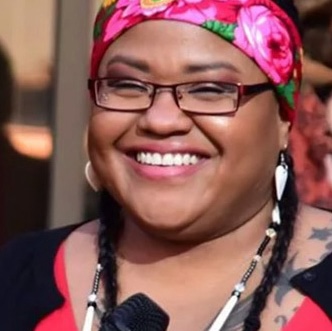 Headshot of tai simpson. She wears a pink floral scarf with her hair in two braids. She is smiling with a microphone in hand.